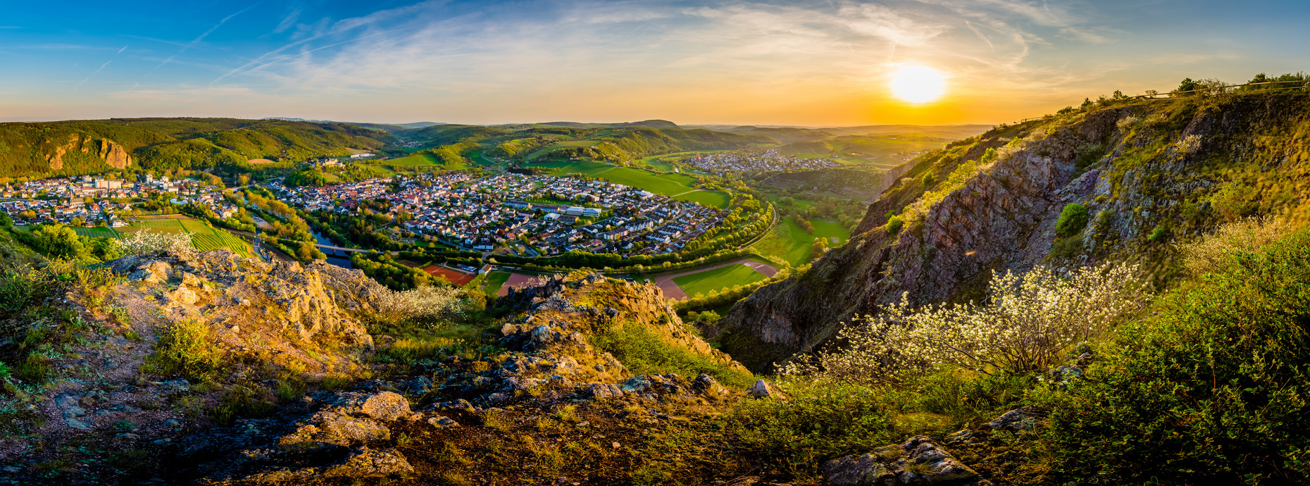 Panoramaaufnahme eines sonnigen Tals mit Kleinstadt, umgeben von grünen Hügeln und Felsen im Sonnenuntergang
